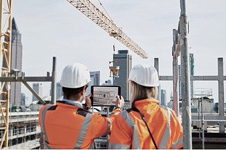 Two people wearing helmets and orange vests inspect a plan on a pad in front of a construction site with a crane