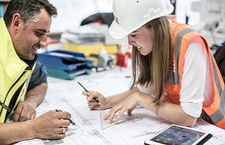 A man in a yellow vest and a woman in a helmet and orange vest work together on a plan, a pad on the table