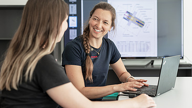 Smiling woman at a laptop in front of a large screen with building plans in the background