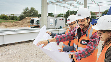 Blick auf einen Plan vor dem drei Personen mit Bauhelmen befinden und darüber sprechen
