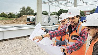View of a plan in front of which three people with construction helmets are standing and talking about it