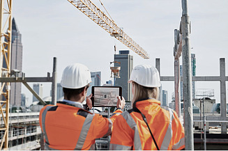Zwei Personen mit Helm und orangefarbener Weste begutachten einen Plan auf einem Pad vor einer Baustelle mit Kran