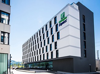 Hvid facade med jævnt fordelte vinduer, glasfronter i stueetagen. To buske i rektangulære plantekasser foran indgangen