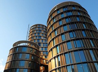 Four staggered round towers with differently sized illuminated windows, with trees and a few passers-by in front of them