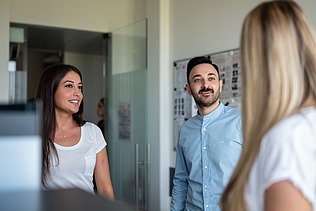 Eine blonde Frau von hinten, ein Mann und eine Frau frontal sprechen miteinander vor einer Metaplanwand