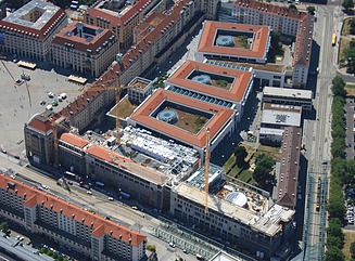 Fireetagers hjørnebygning i wilhelminsk stil med arkade på en stor plads med biler og fodgængere i forgrunden